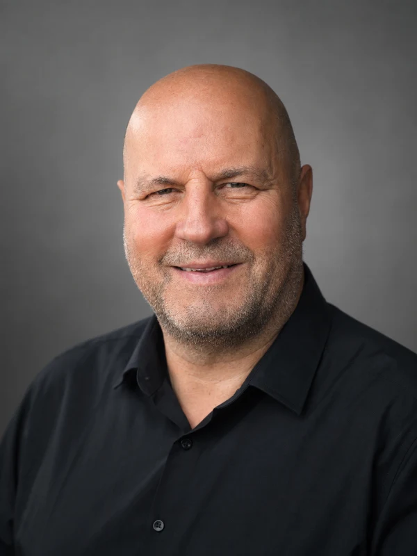 Mike Hesse, lächelnd, trägt schwarzes Polohemd vor grauem Hintergrund.